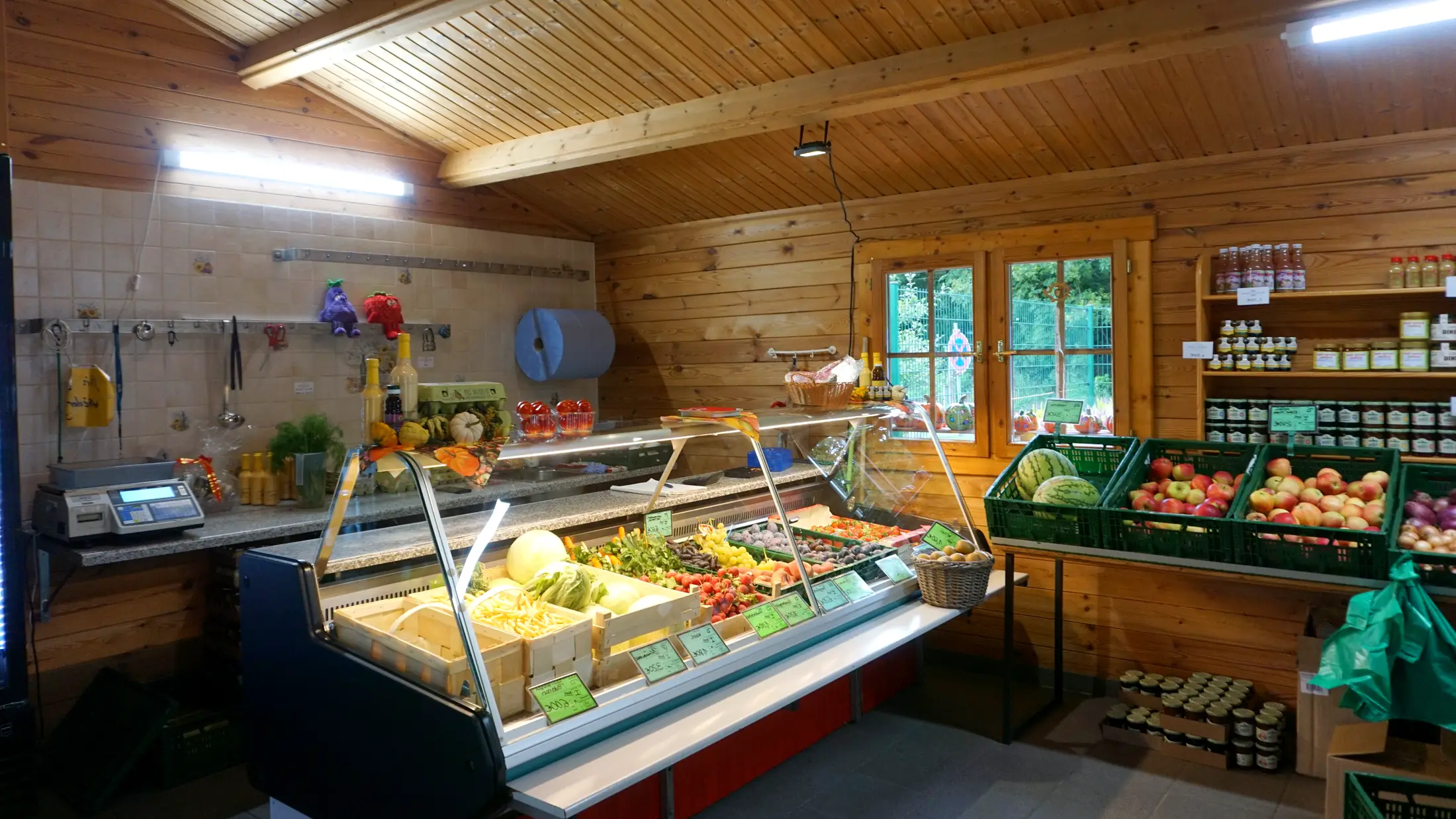 Blick in den Hofladen: Kühltheke mit frischem Obst und Gemüse, daneben Kisten mit Äpfeln und Melonen